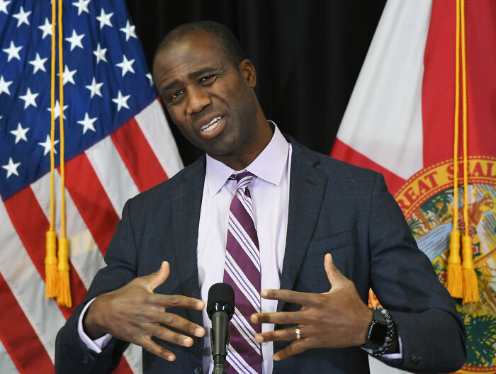 Man in suit speaking at podium with American and Florida state flags in background, discussing CDC vaccine plans controversy.