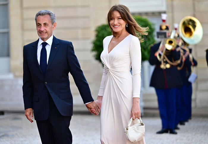 Former French President Nicolas Sarkozy holding hands with a woman outdoors, with musicians playing brass instruments in the background.