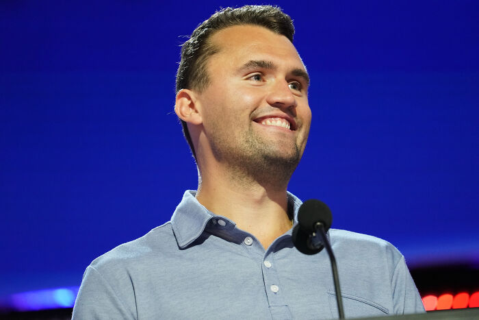 Man smiling and speaking at a microphone during event with blue background, related to Charlie Kirk GOP currency proposal.