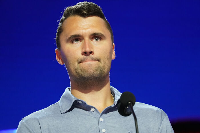 Young man speaking at a podium with a microphone against a blue background, related to Charlie Kirk assassination topic.