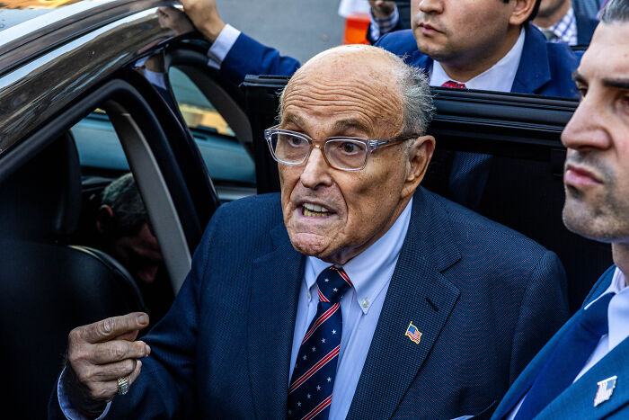 Rudy Giuliani wearing a suit with an American flag pin, speaking near a car surrounded by men in formal attire.