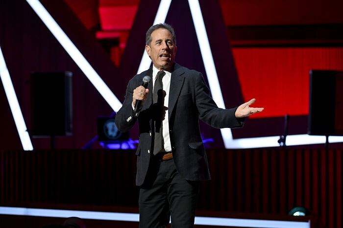 Comedian Jerry Seinfeld performing on stage with microphone, discussing Free Palestine movement and hate group comparison.