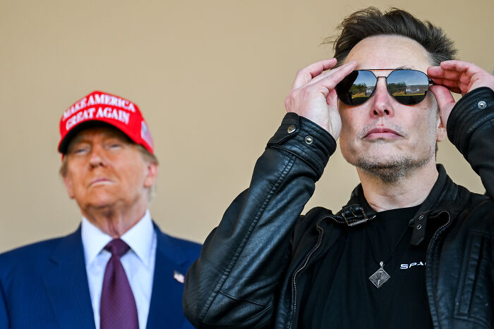 Donald Trump and Elon Musk pictured together, with Trump wearing a red hat and Musk adjusting sunglasses outdoors.