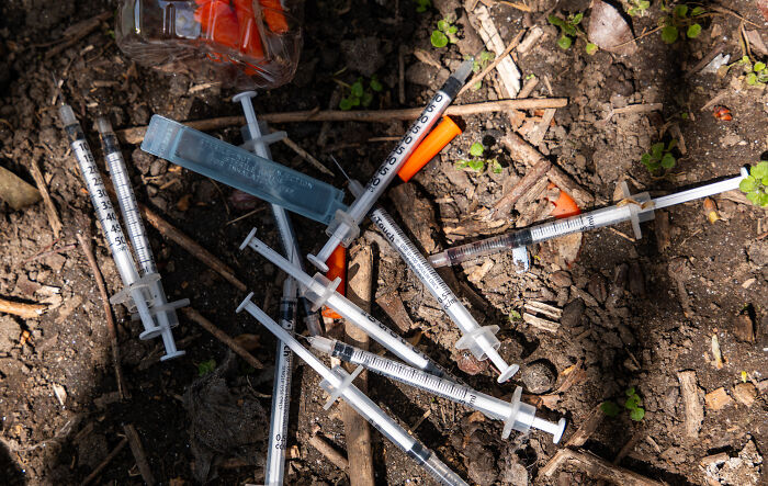 Discarded hypodermic syringes and a small container scattered on dirt, highlighting illegal substances impact in America.