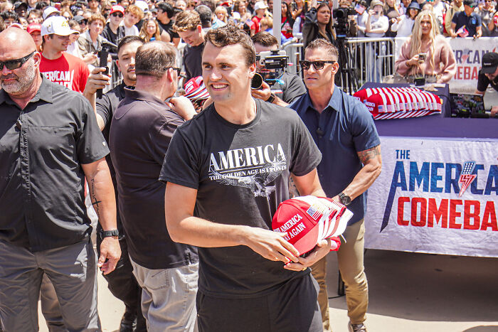 Man holding red hats at a crowded rally highlighting concerns about America&rsquo;s escalating political violence.