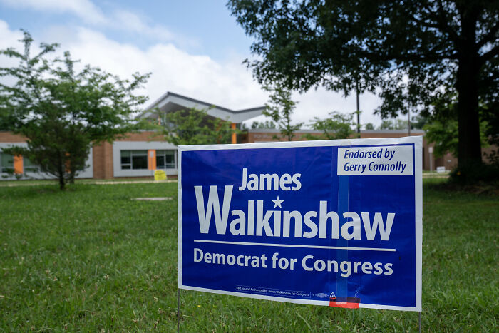 Blue campaign sign for Democrat James Walkinshaw for Congress displayed on grass with trees and a modern building in background.