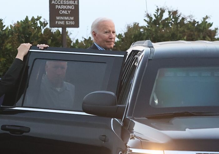 Joe Biden with a visible head wound getting into a black vehicle near an accessible parking sign outdoors.