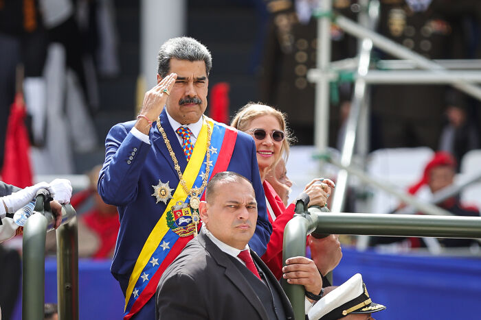 Nicolas Maduro wearing presidential sash saluting during public event with security and attendees, hinting at Venezuela conflict concerns.