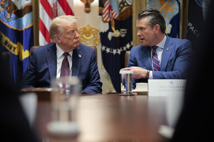 Donald Trump in a suit talking with a man about renaming Department of Defense to Department of War in a formal meeting room.