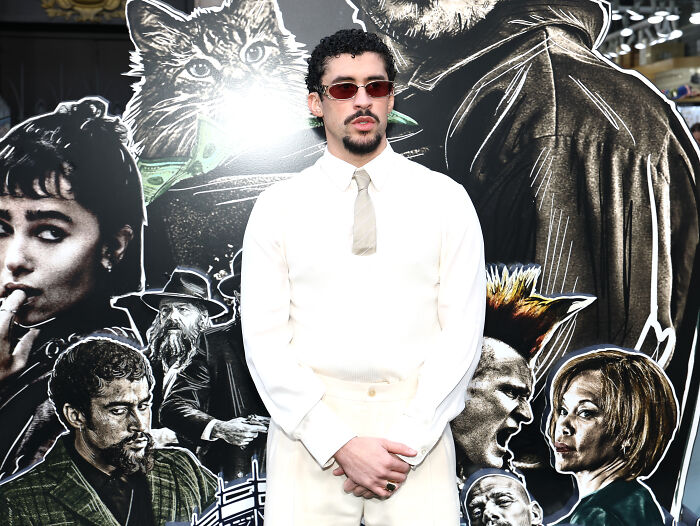 Bad Bunny wearing sunglasses and white outfit, standing in front of artistic character cutouts at an event.