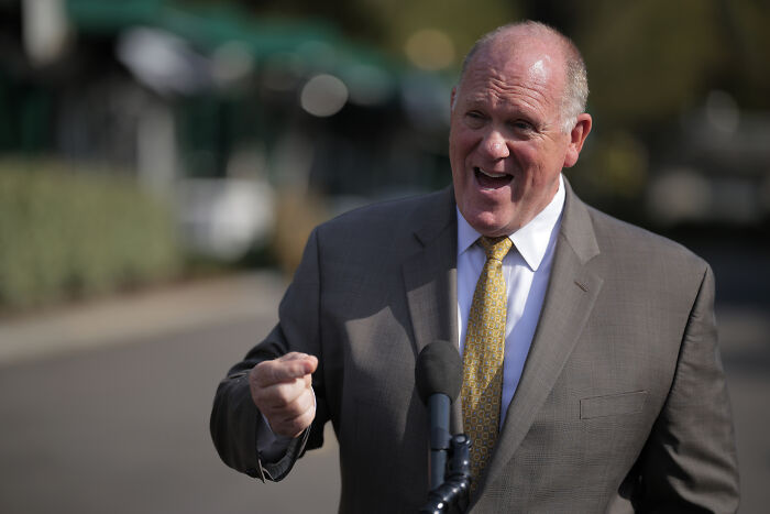 Tom Homan speaking outdoors near a microphone, wearing a suit and yellow tie in a sunny setting.