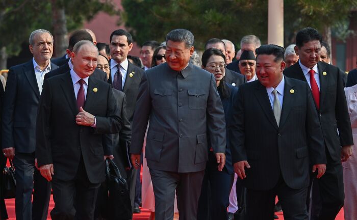 Leaders Putin, Xi, and Kim walking together during a formal event, highlighting claims of conspiracy against the U.S.