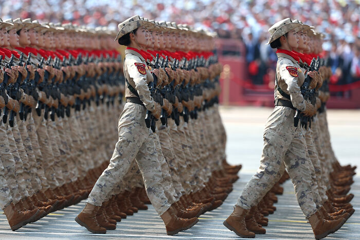 Female soldiers in camouflage uniforms marching in formation during a military parade related to conspiracy claims against the U.S.