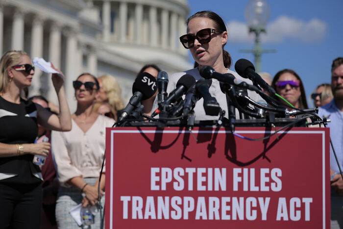 Woman wearing sunglasses speaking at a podium about Epstein files transparency act during a tearful Epstein victims rally.