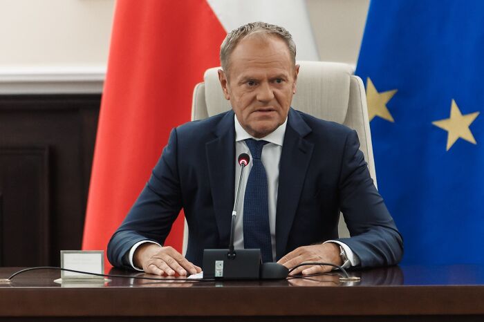 Polish official in a suit speaking at a press conference with Poland and European Union flags behind him.
