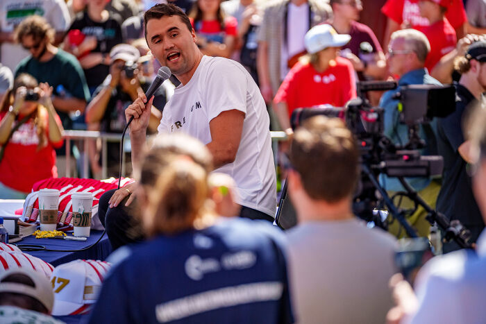Man speaking into a microphone at an outdoor event with a crowd, linked to left-wing hashtag activists and Charlie Kirk.