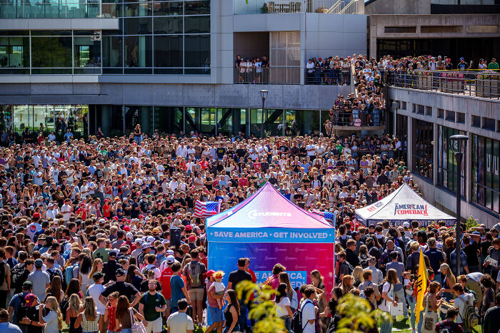 Large crowd gathered at an outdoor event with tents displaying Save America and American Comeback messages, reflecting political activism.