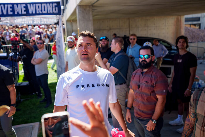 Man wearing a white freedom shirt at a crowded outdoor event with people and security in the background