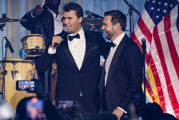 Two men in tuxedos speaking on stage with microphones, an American flag and musical instruments in the background.