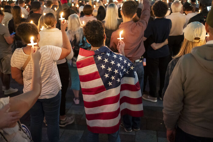 Crowd holding candles with one person draped in American flag highlighting America&rsquo;s escalating political violence concerns.