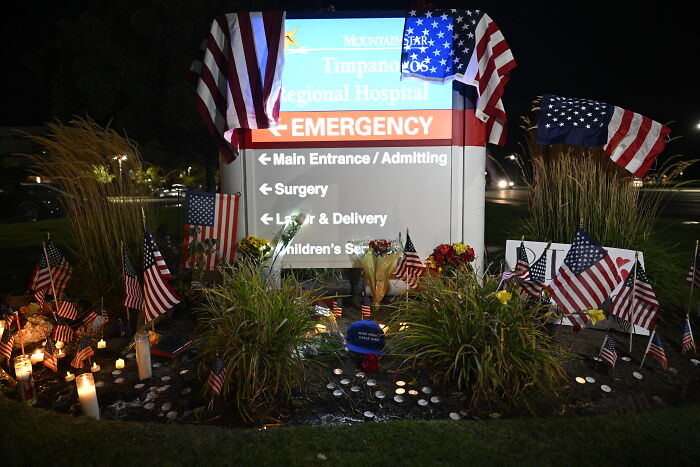 Memorial with American flags, candles, and flowers in front of hospital emergency sign reflecting firearm fatalities impact.