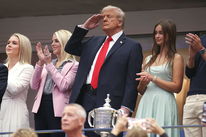Donald Trump standing and saluting at an event, with people applauding around him and a trophy in the foreground.