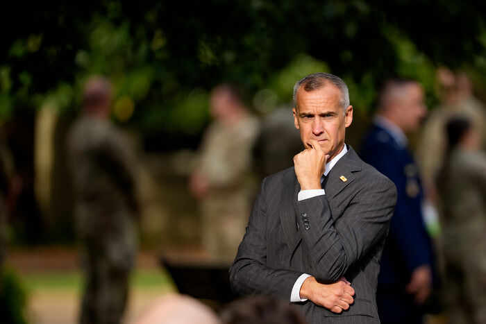 Man in a gray suit standing thoughtfully outdoors with blurred figures in the background during a political event in D.C.