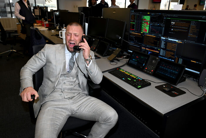 Conor McGregor in a grey suit talking on phone in a trading office with multiple computer screens around him