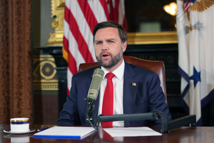 Man in suit with red tie speaking into microphone in formal room, discussing Utah quake and Charlie Kirk assassination claims.