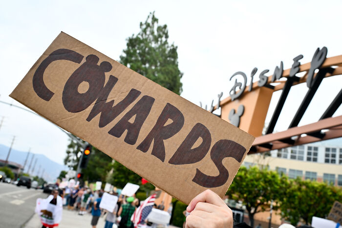 Protesters holding a cardboard sign reading cowards outside Walt Disney Studios amid Jimmy Kimmel suspension and ratings boost news.
