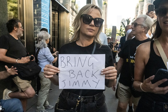 Woman wearing sunglasses holds bring back Jimmy sign during street protest related to Jimmy Kimmel returning to ABC sources say