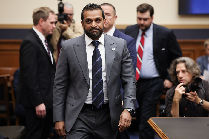 Man in gray suit and striped tie walking in hearing room with people and photographers behind him, FBI Director Kash Patel context