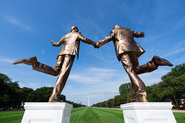 Bronze Trump and Epstein statue holding hands, standing on white pedestals with a clear blue sky and Washington Monument background.