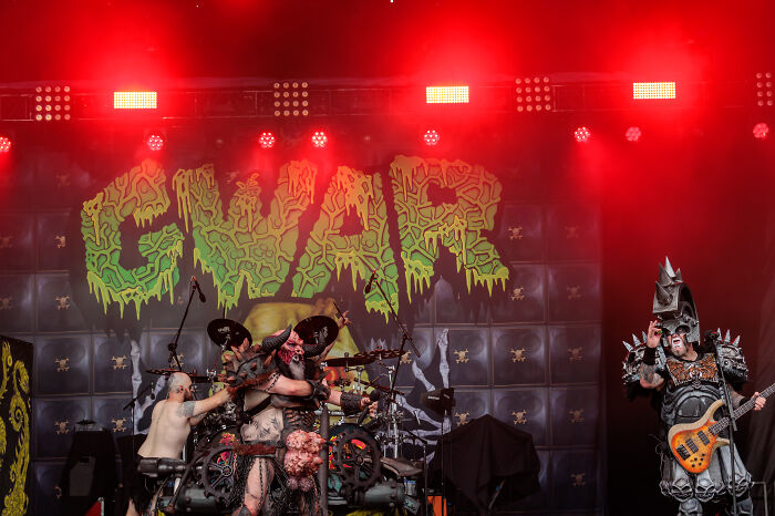 Rock band performing onstage with MAGA-themed costumes and props under red stage lighting at a live concert.