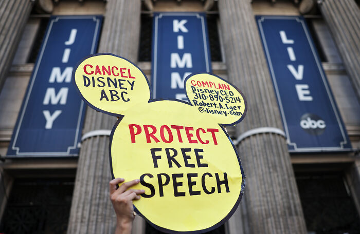 Protester holding a sign to protect free speech with Jimmy Kimmel Live banners on a building in the background.