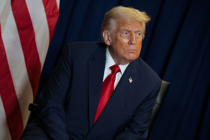 Donald Trump sitting in a dark suit and red tie with a serious expression, against an American flag backdrop.