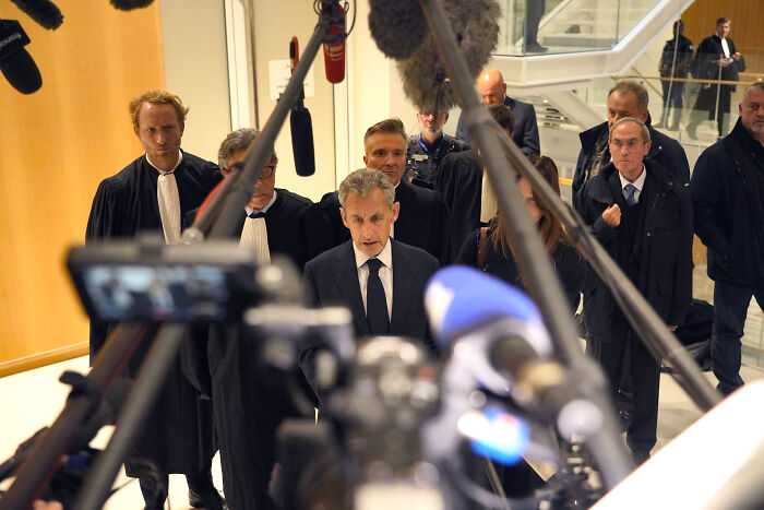 Former French President Nicolas Sarkozy speaking to media while surrounded by legal team and reporters inside a courthouse.
