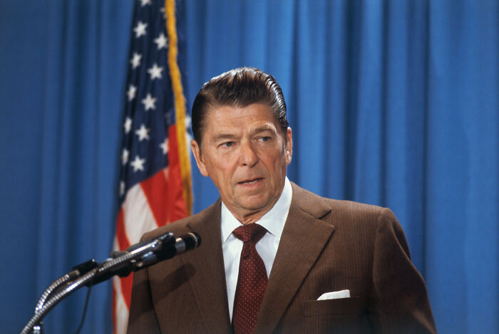 American president in a brown suit speaking at a podium with an American flag and blue curtain backdrop, representing war leaders.
