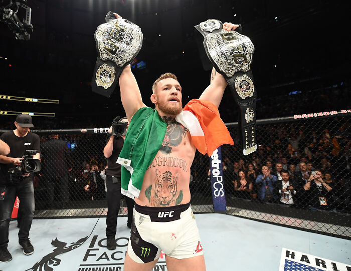 Conor McGregor inside UFC octagon holding two championship belts with Irish flag draped over his shoulders.