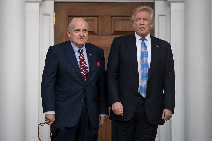 Rudy Giuliani and Donald Trump standing outdoors near wooden door, discussing presidential medal of freedom news.