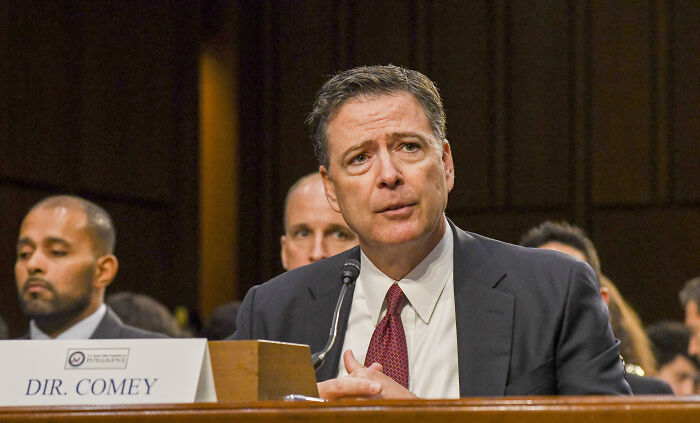Former FBI Chief James Comey speaking at a hearing, wearing a dark suit and red tie, appearing serious and attentive.