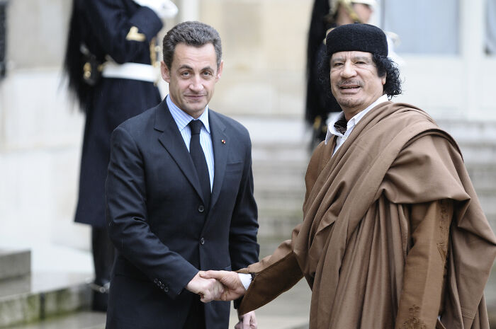 Former French President Nicolas Sarkozy shaking hands with another man outdoors, dressed formally in a suit.