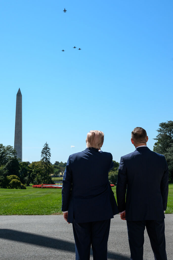 Trump and another man in suits watch a thunderous jet flyover near the Washington Monument on a clear day.