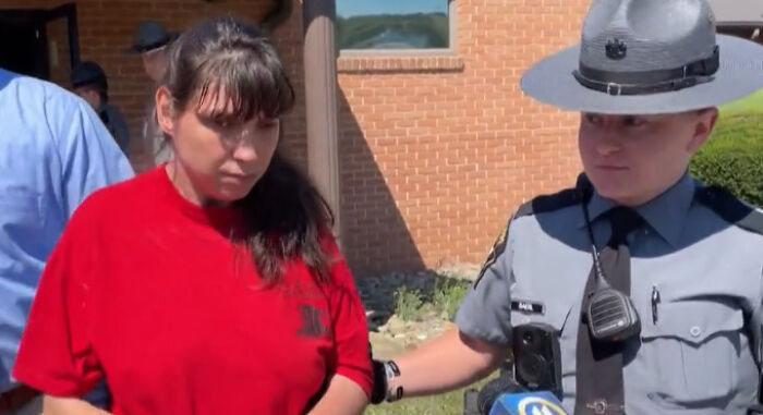 Woman in red shirt being escorted by police officer outside a brick building after children found in dungeon conditions