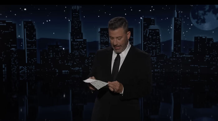 Jimmy Kimmel on a late-night set wearing a black suit, reading notes while mocking Trump in his monologue.