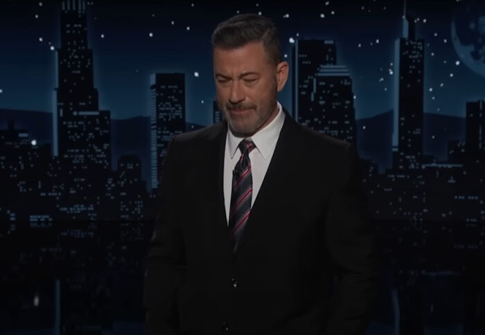 Jimmy Kimmel on late-night set, wearing suit and tie, during a monologue on a nighttime cityscape backdrop.