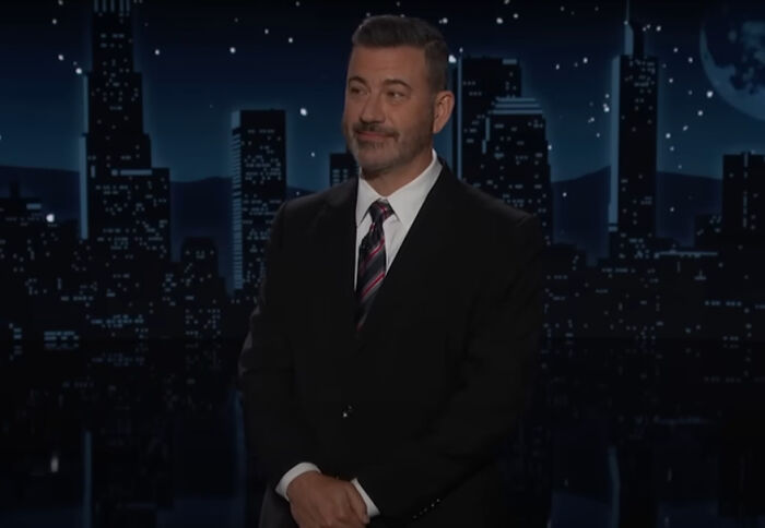 Jimmy Kimmel in a dark suit standing on a late-night show set with city skyline background at night
