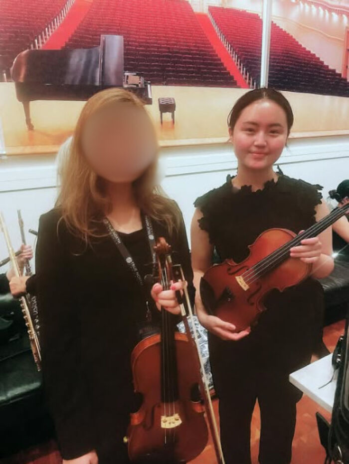 Teenage musician holding violin with another musician blurred, posing in concert hall after performance.