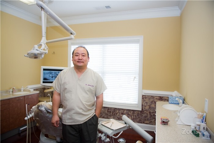 Dentist standing in exam room of his clinic with dental equipment and bright window behind him.
