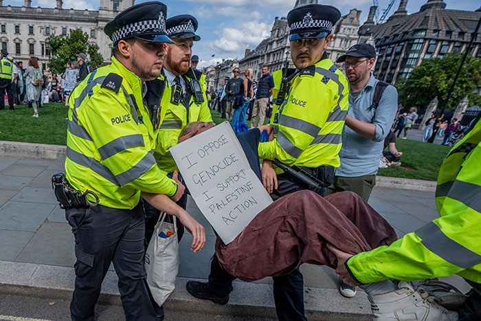 UK police officers detaining protester holding sign amid free speech and social media laws debate in the UK.
